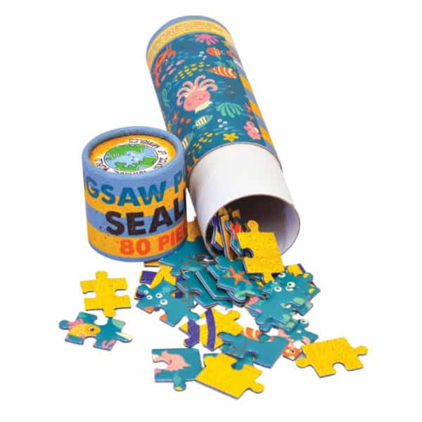 Blue and yellow puzzle pieces spill out of a carbourd cylinder decorated in a sealife scene. Lid says Jigsaw Puzzle Sealife 80 pieces